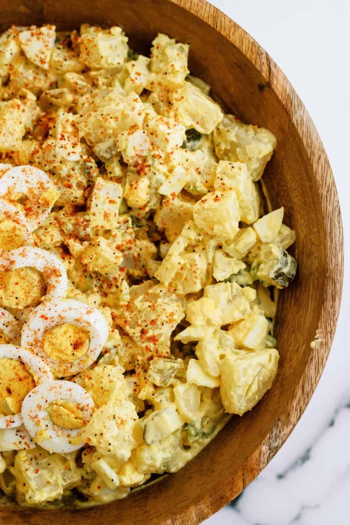 A wooden bowl filled with potato salad topped with sliced hard-boiled eggs and sprinkled with paprika.
