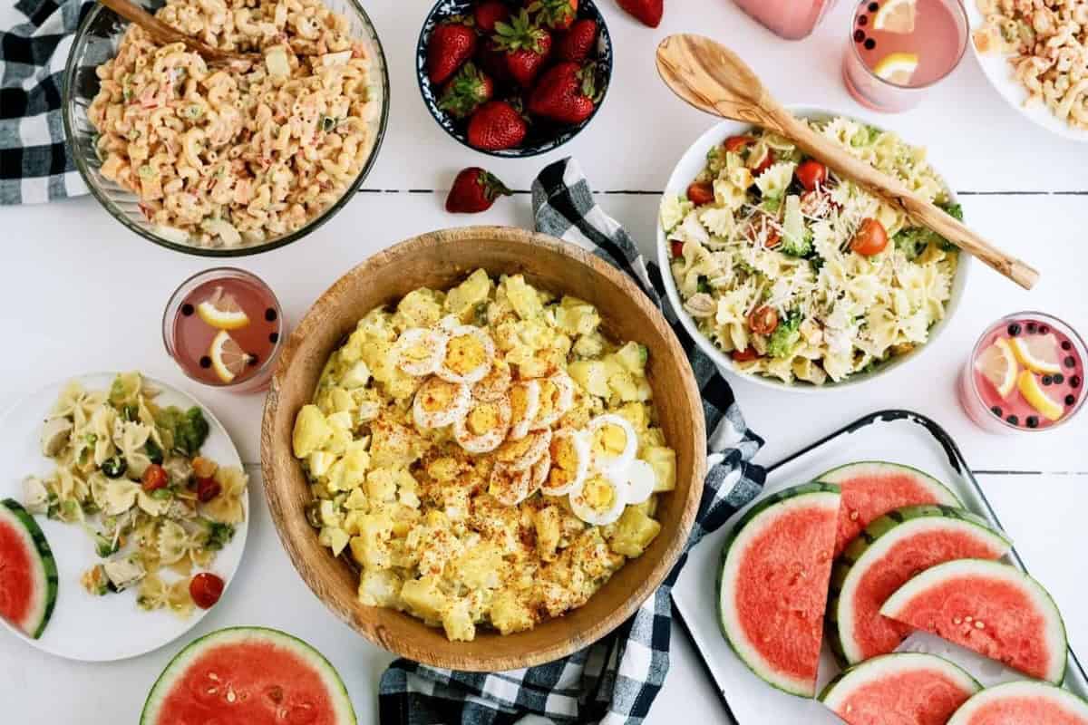 A table with dishes including potato salad with boiled eggs, pasta salad, watermelon slices, strawberry bowl, drinks with lemon slices, and a checkered napkin.