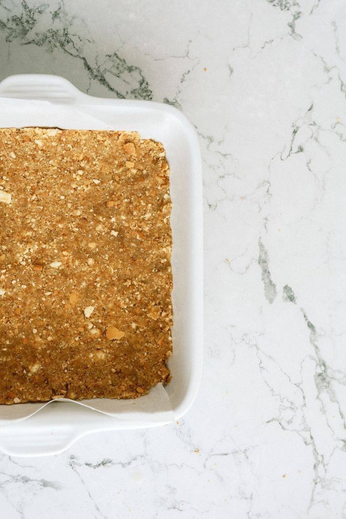 A white baking dish with a pressed, crumbly crust mixture inside, placed on a white marble countertop.