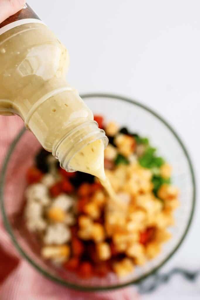 A hand pours creamy dressing from a bottle over a salad with chopped vegetables in a glass bowl.