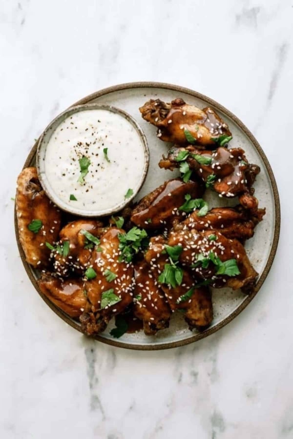 A plate of glazed chicken wings with sesame seeds and cilantro, accompanied by a bowl of creamy dipping sauce, on a marble surface.