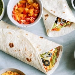 Two burritos filled with vegetables and olives on a table, surrounded by bowls of chopped tomatoes and shredded cheddar cheese.