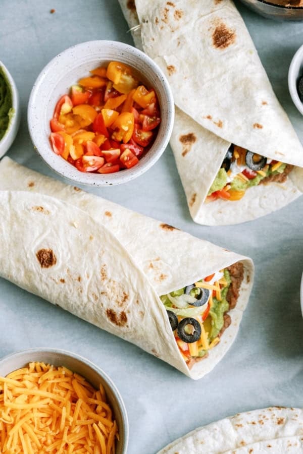 Two burritos filled with vegetables and olives on a table, surrounded by bowls of chopped tomatoes and shredded cheddar cheese.