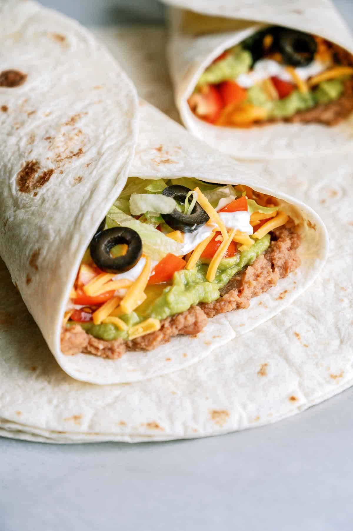 A close-up of a burrito filled with refried beans, guacamole, shredded cheese, lettuce, tomatoes, and sliced black olives, wrapped in a flour tortilla.