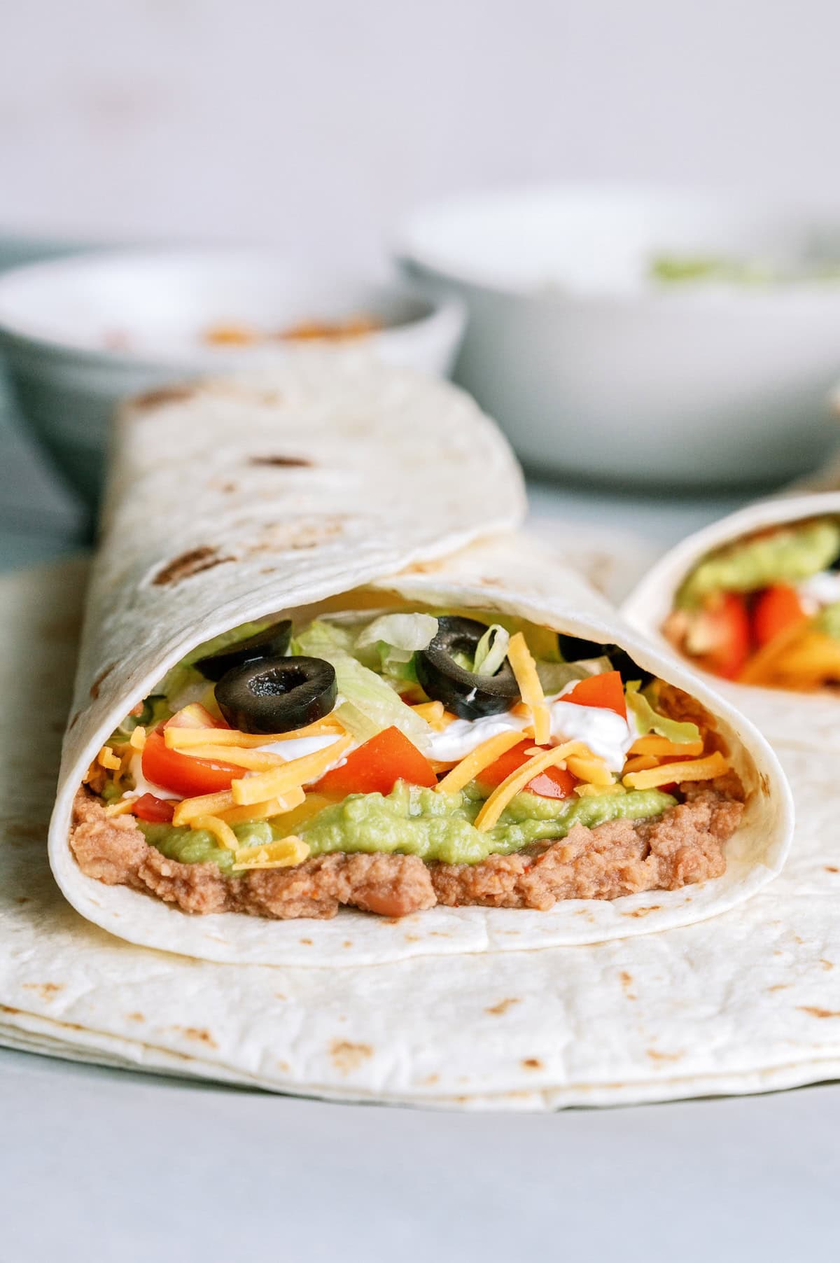 A burrito with refried beans, guacamole, shredded cheese, lettuce, sour cream, black olives, and diced red peppers on a flour tortilla.