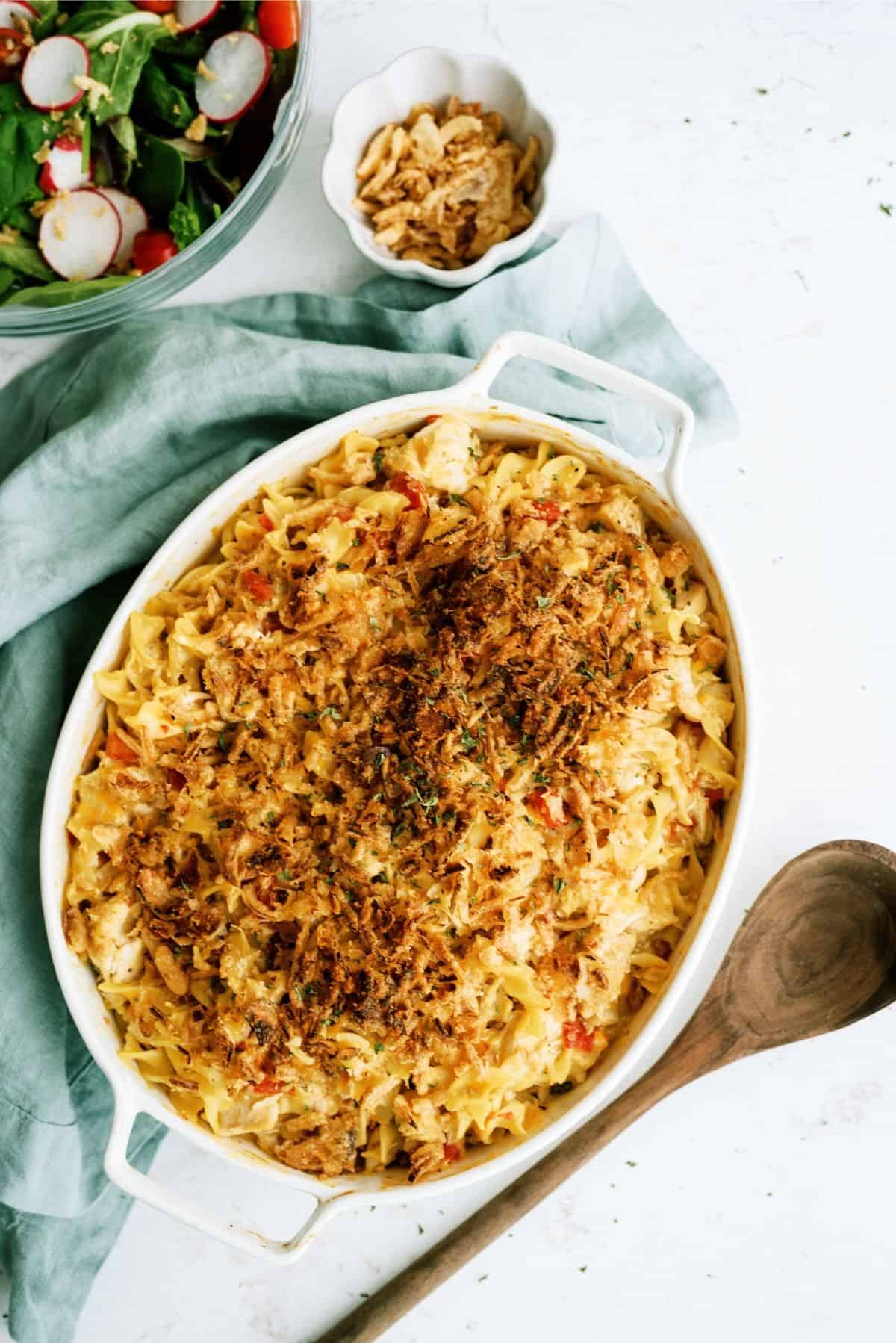 French Onion Chicken Noodle Casserole in a white casserole dish with a wooden spoon and bowl of side salad