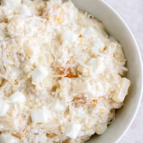 A white bowl filled with a creamy mixture containing marshmallows, shredded coconut, pineapple, and other fruit pieces.