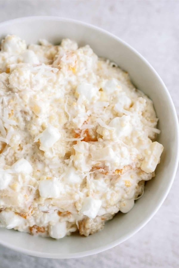 A white bowl filled with a creamy mixture containing marshmallows, shredded coconut, pineapple, and other fruit pieces.