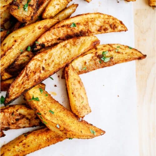 Seasoned potato wedges garnished with parsley on a sheet of parchment paper on a wooden surface.