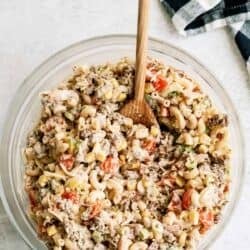 A bowl of macaroni salad with ground meat, chopped tomatoes, and vegetables, with a wooden spoon. A checkered cloth is nearby on a light-colored surface.