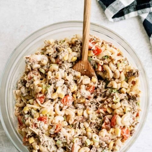 A bowl of macaroni salad with ground meat, chopped tomatoes, and vegetables, with a wooden spoon. A checkered cloth is nearby on a light-colored surface.