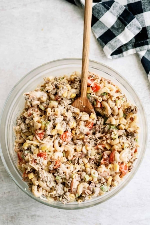 A bowl of macaroni salad with ground meat, chopped tomatoes, and vegetables, with a wooden spoon. A checkered cloth is nearby on a light-colored surface.