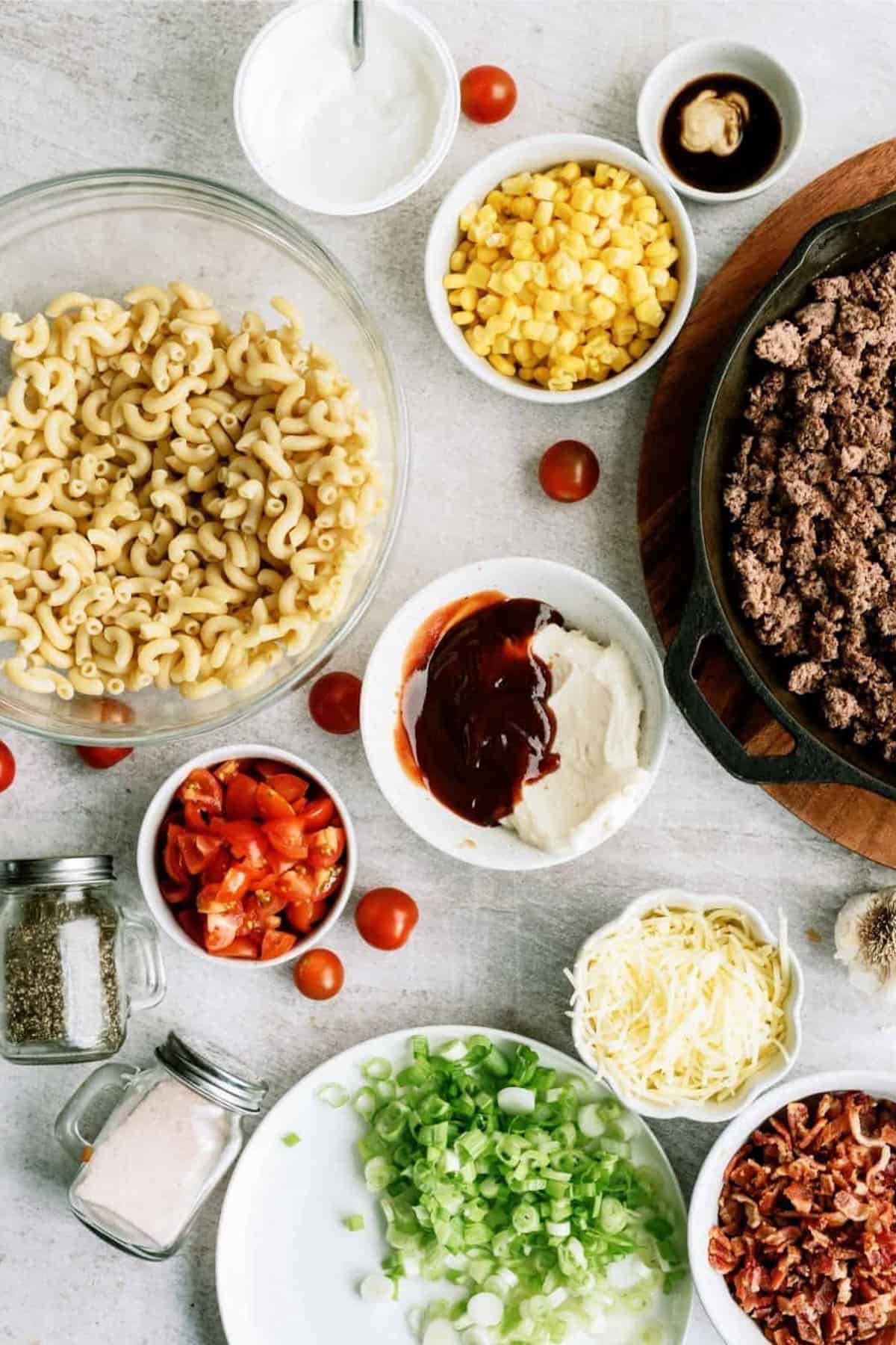 A variety of ingredients for a taco pasta salad, including cooked pasta, ground beef, corn, tomatoes, green onions, shredded cheese, bacon, sour cream, and seasonings, arranged on a table.