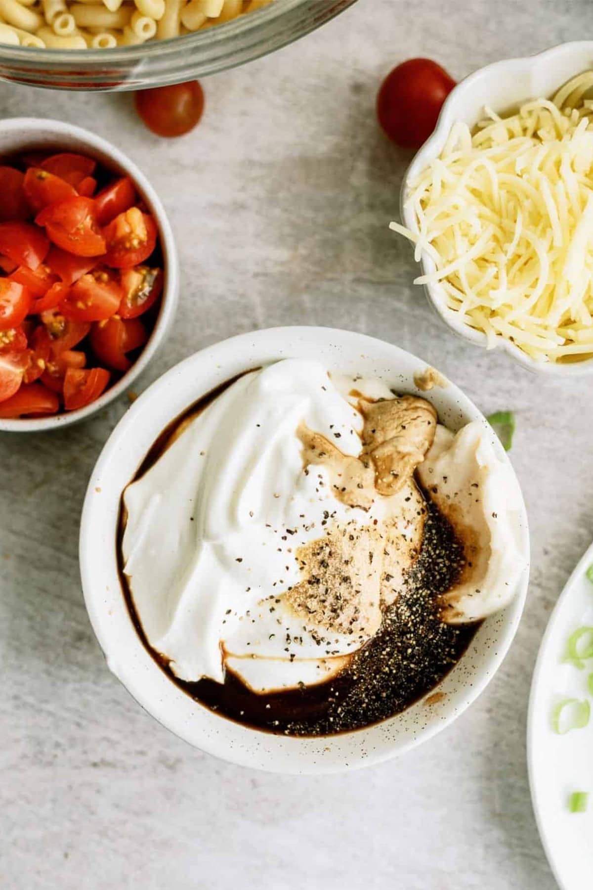 A bowl with yogurt topped with black pepper and sauce, surrounded by chopped tomatoes, grated cheese, green onions, and pasta on a gray surface.