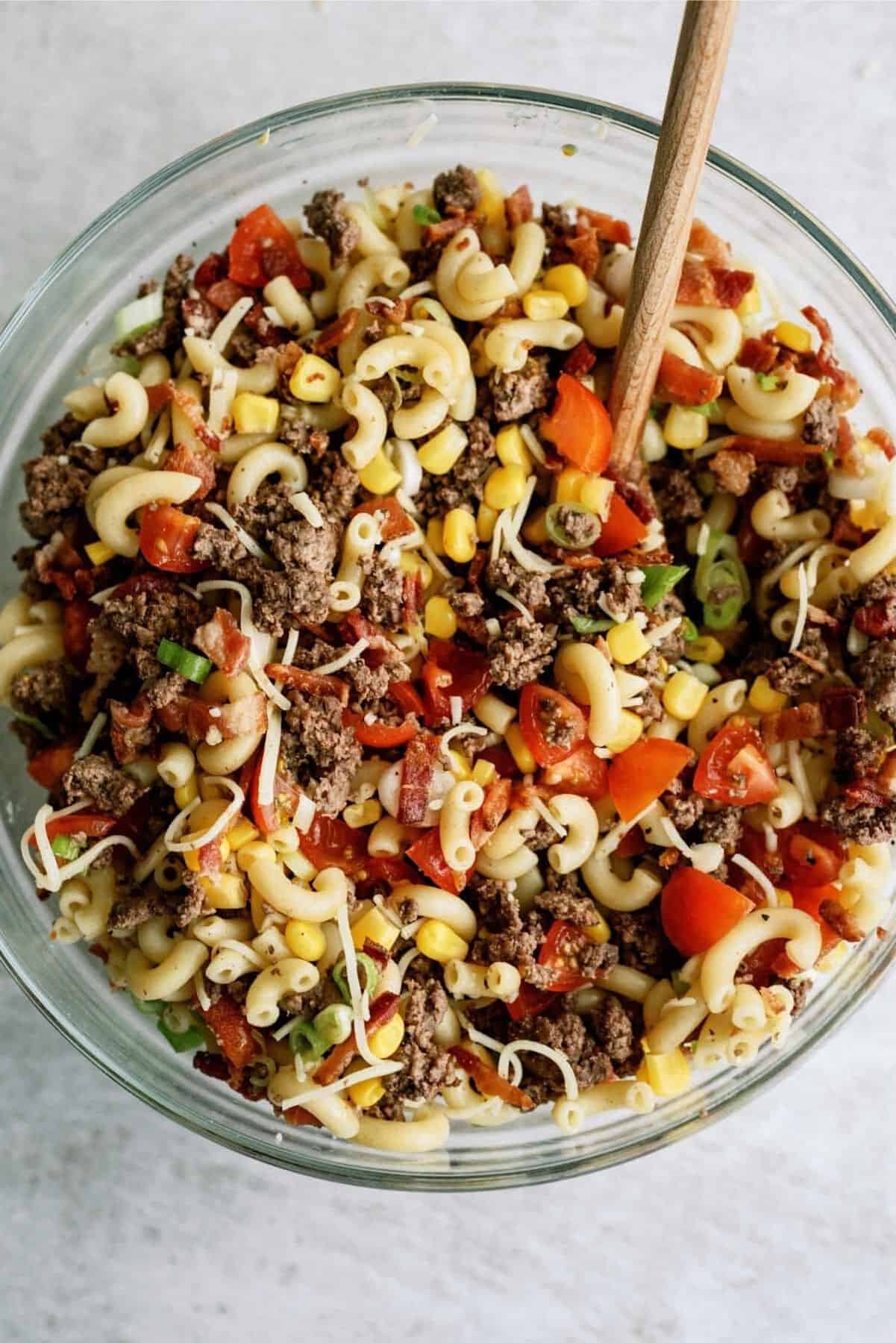 A glass bowl filled with a mixture of macaroni, ground beef, diced tomatoes, corn, and green onions, stirred with a wooden spoon.