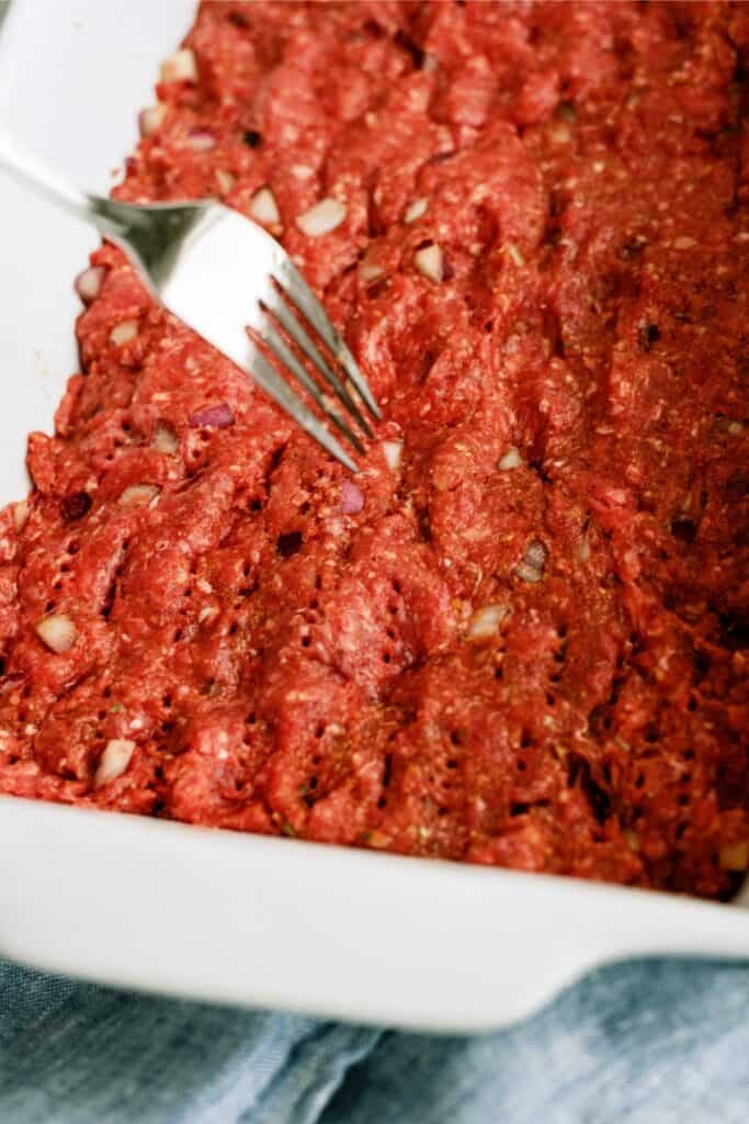 A fork poking holes in the beef mixture that is pressed into the baking dish.