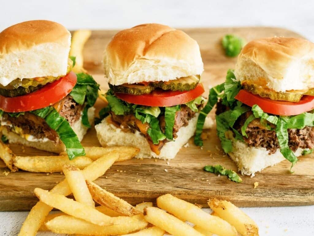 3 baked cheeseburger sliders in a row on a cutting board with french fries in front of them.