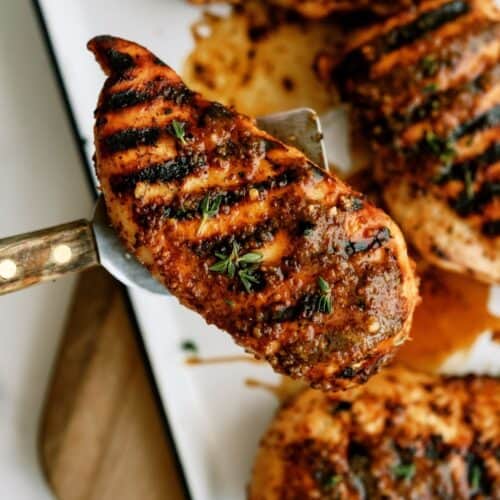 Grilled chicken breast with char marks, seasoned with herbs, is lifted from a tray with a metal spatula. Other pieces are visible in the background.
