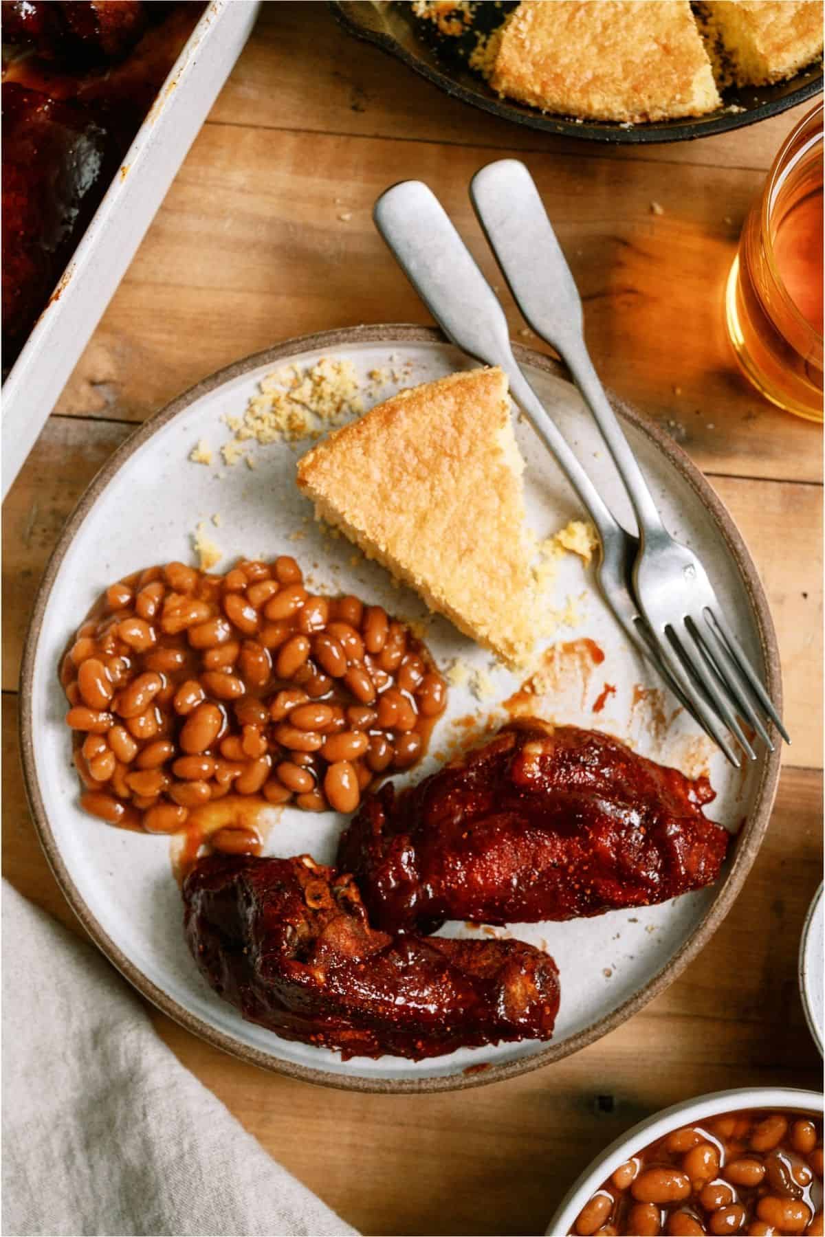 A plate with baked beans, two pieces of barbecued ribs, and a slice of cornbread, with a fork and knife beside the food on a wooden table.