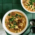 Two bowls of beef noodle soup with parsley garnish on a green tiled surface, accompanied by two black spoons.