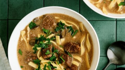 Two bowls of beef noodle soup with parsley garnish on a green tiled surface, accompanied by two black spoons.