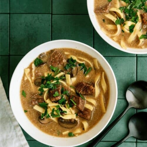 Two bowls of beef noodle soup with parsley garnish on a green tiled surface, accompanied by two black spoons.