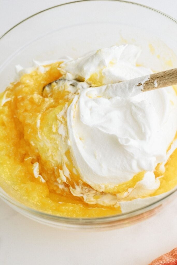 Folding whip cream into a fruit mixture in a glass bowl.