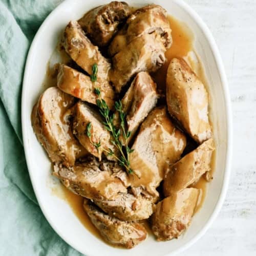 Sliced pork tenderloin with brown gravy, garnished with fresh thyme, served on a white oval plate beside a green cloth.
