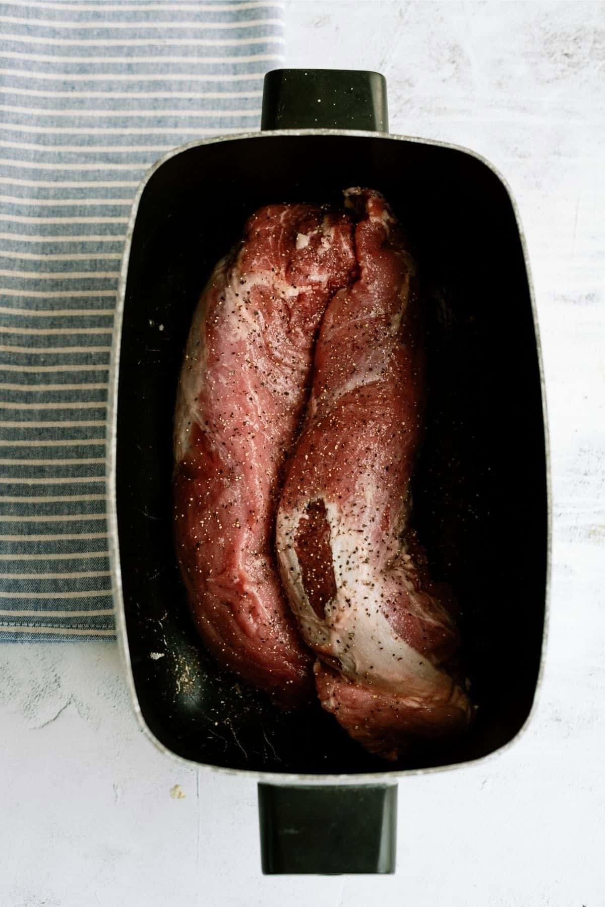 Two raw pork tenderloins seasoned with pepper in a black roasting pan, placed on a light surface next to a striped cloth.