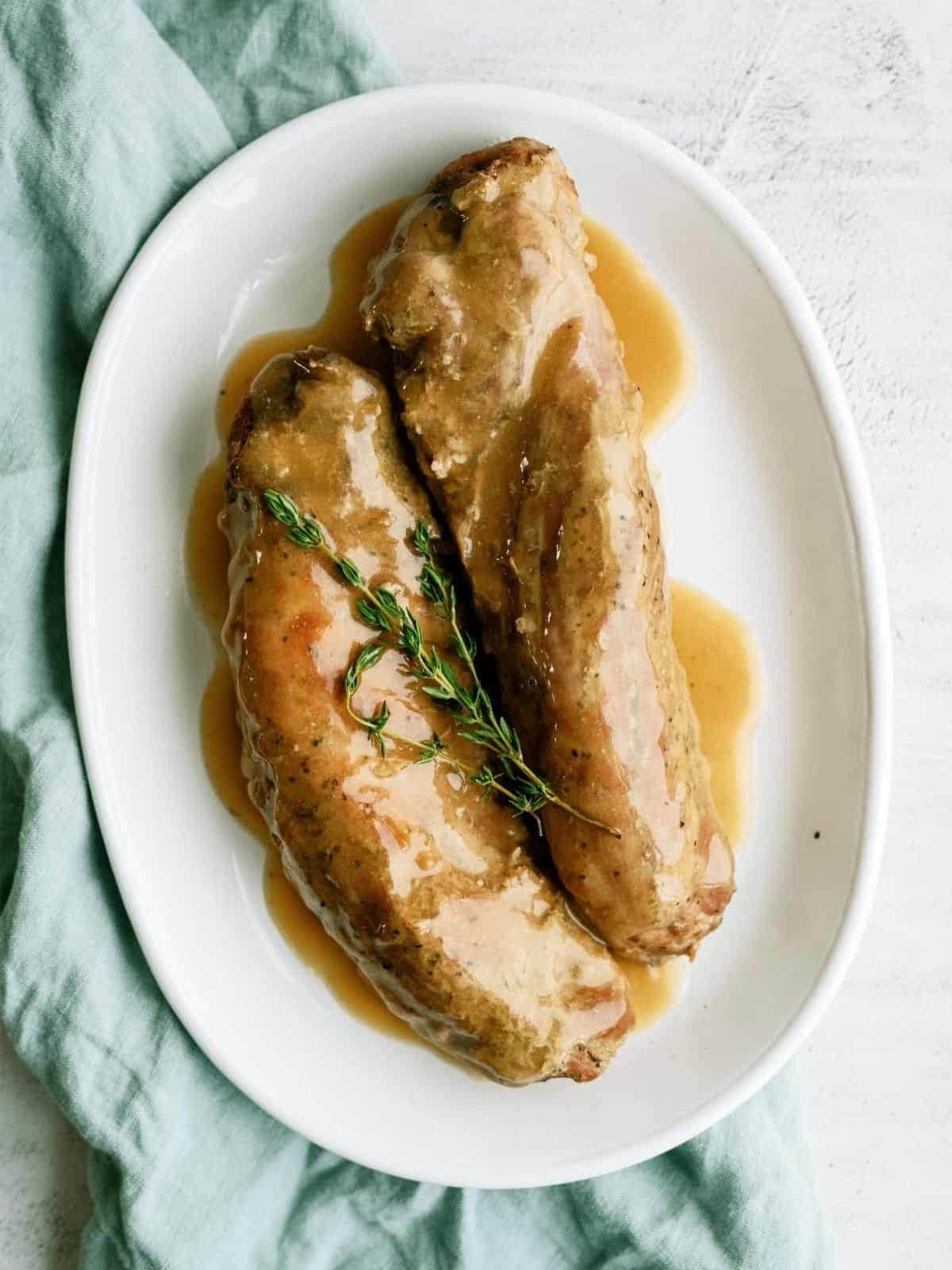 Two pieces of pork tenderloin with brown gravy and a sprig of fresh thyme on a white oval plate, placed on a light green cloth.