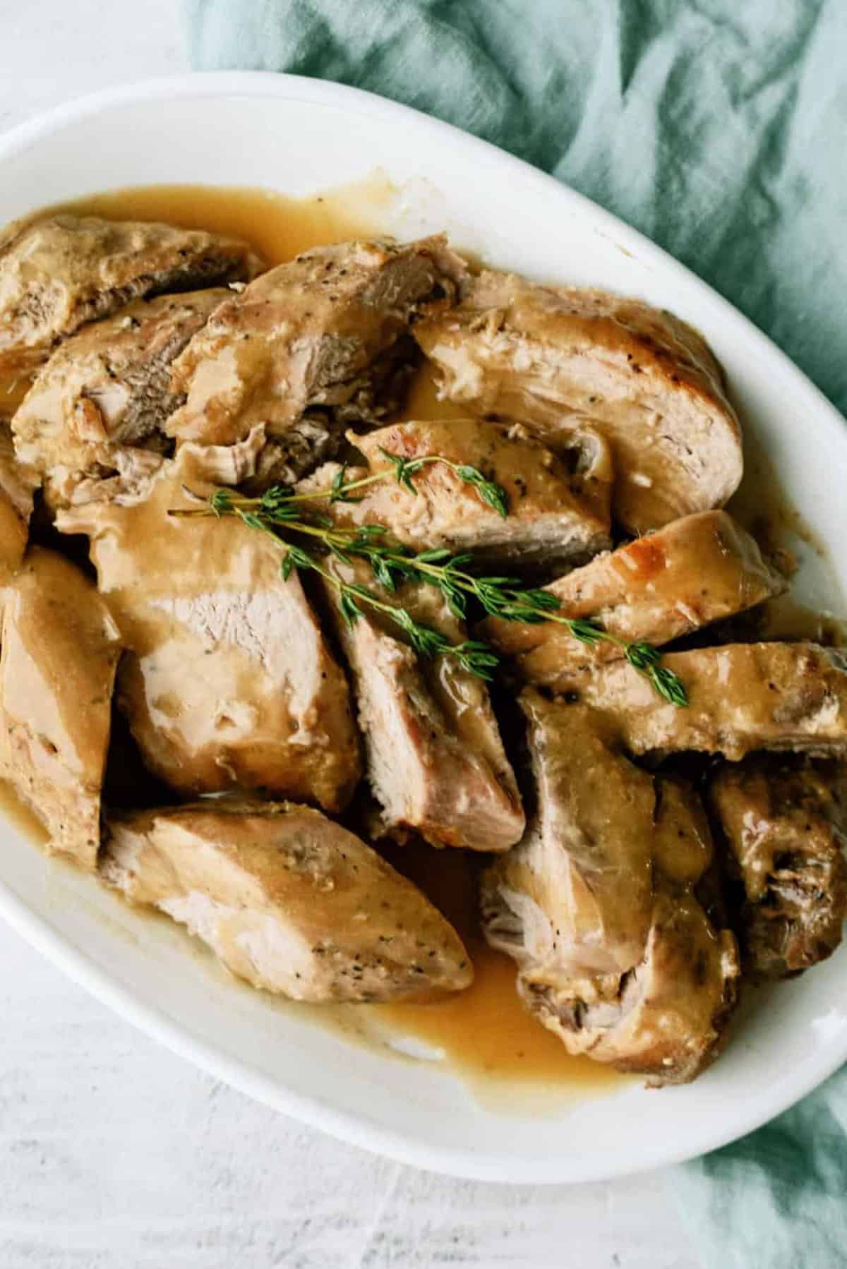 Sliced cooked turkey breast with gravy on a white oval plate, garnished with fresh thyme, next to a green cloth napkin.
