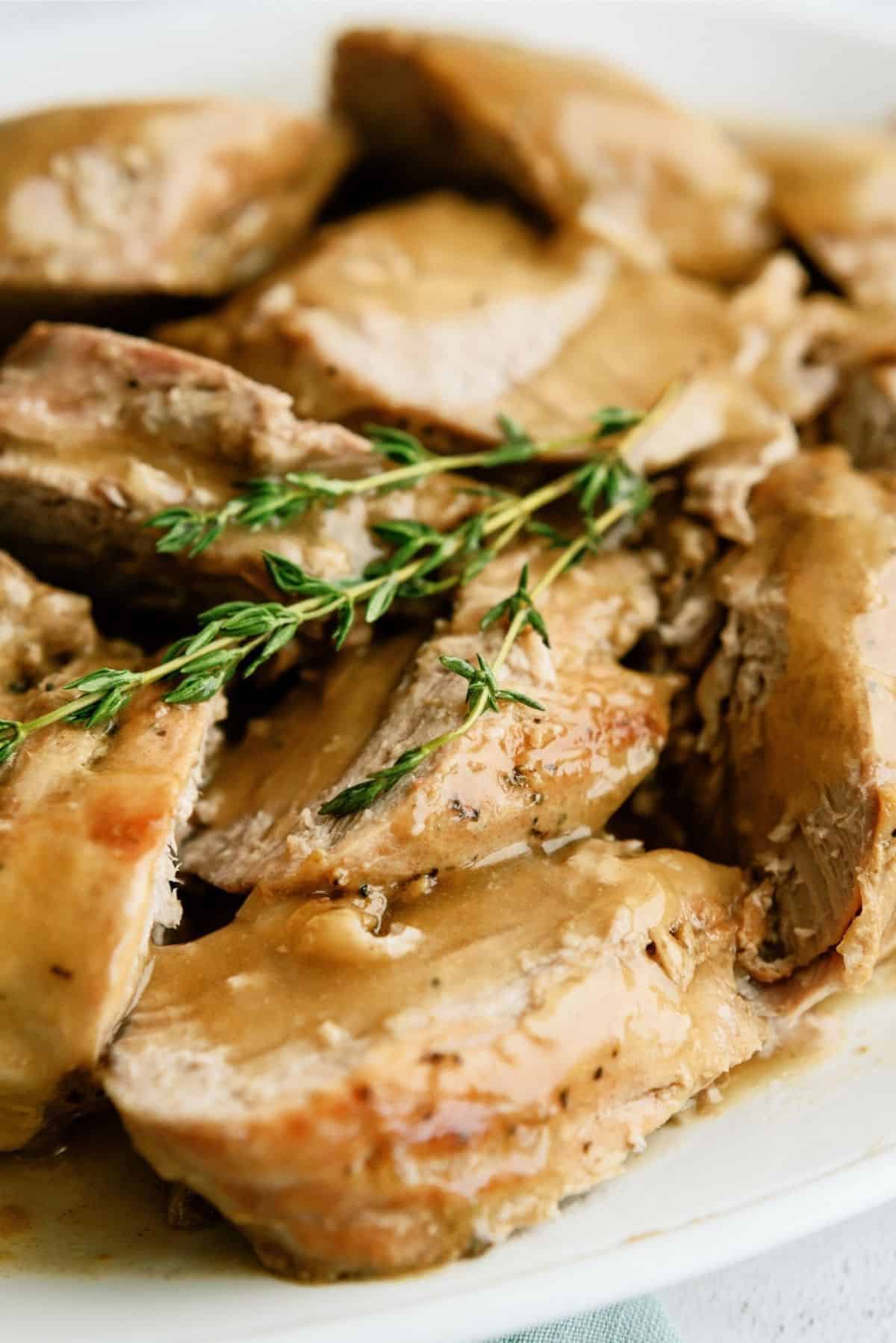 Slices of roast pork topped with brown gravy and garnished with fresh thyme are arranged on a white plate.