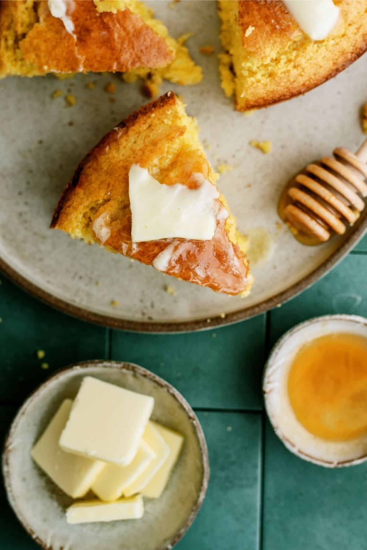Top view of a slice of Easy Homemade Cornbread on a plate with a pat of butter and honey on top of it. Other slices on the plate as well.