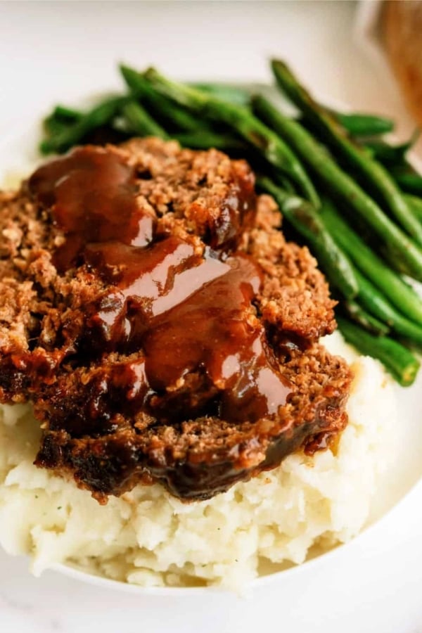 A serving of meatloaf topped with brown gravy, placed on a bed of mashed potatoes, with a side of green beans.