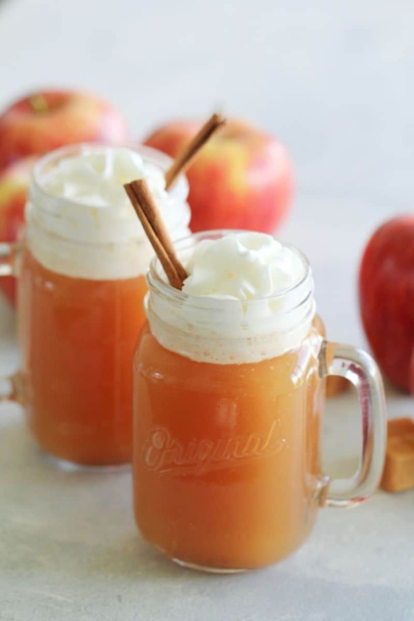 Two glass mugs of apple cider topped with whipped cream and garnished with cinnamon sticks, with apples in the background.