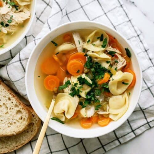 A bowl of chicken tortellini soup with carrots and herbs, placed on a white checkered cloth, next to slices of bread.