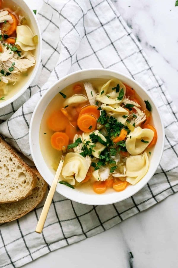 A bowl of chicken tortellini soup with carrots and herbs, placed on a white checkered cloth, next to slices of bread.
