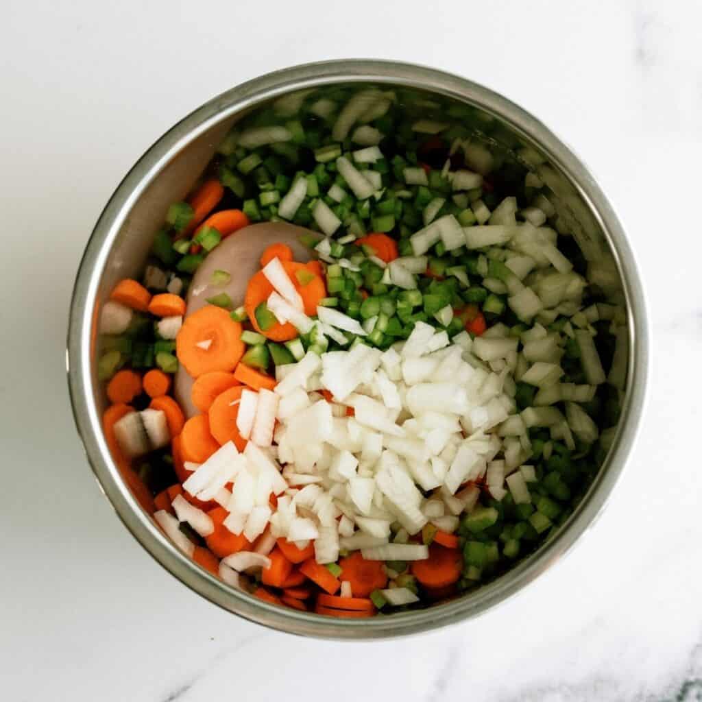A metal bowl containing chopped carrots, celery, onions, and what appears to be raw chicken pieces on a white surface.