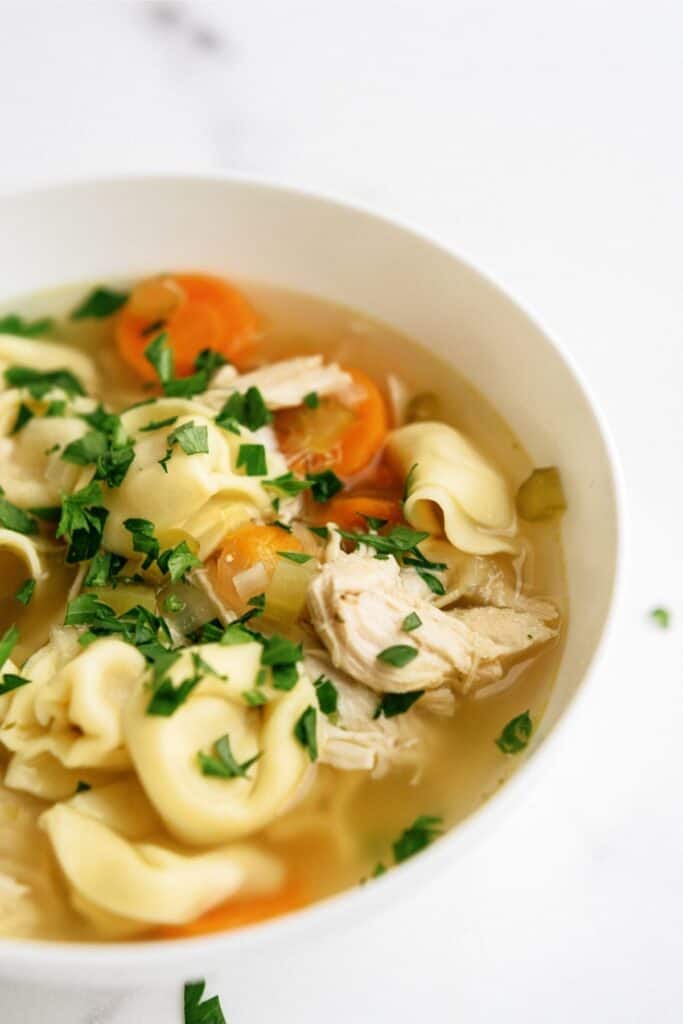 A bowl of chicken tortellini soup with sliced carrots, celery, shredded chicken, and fresh parsley on top.