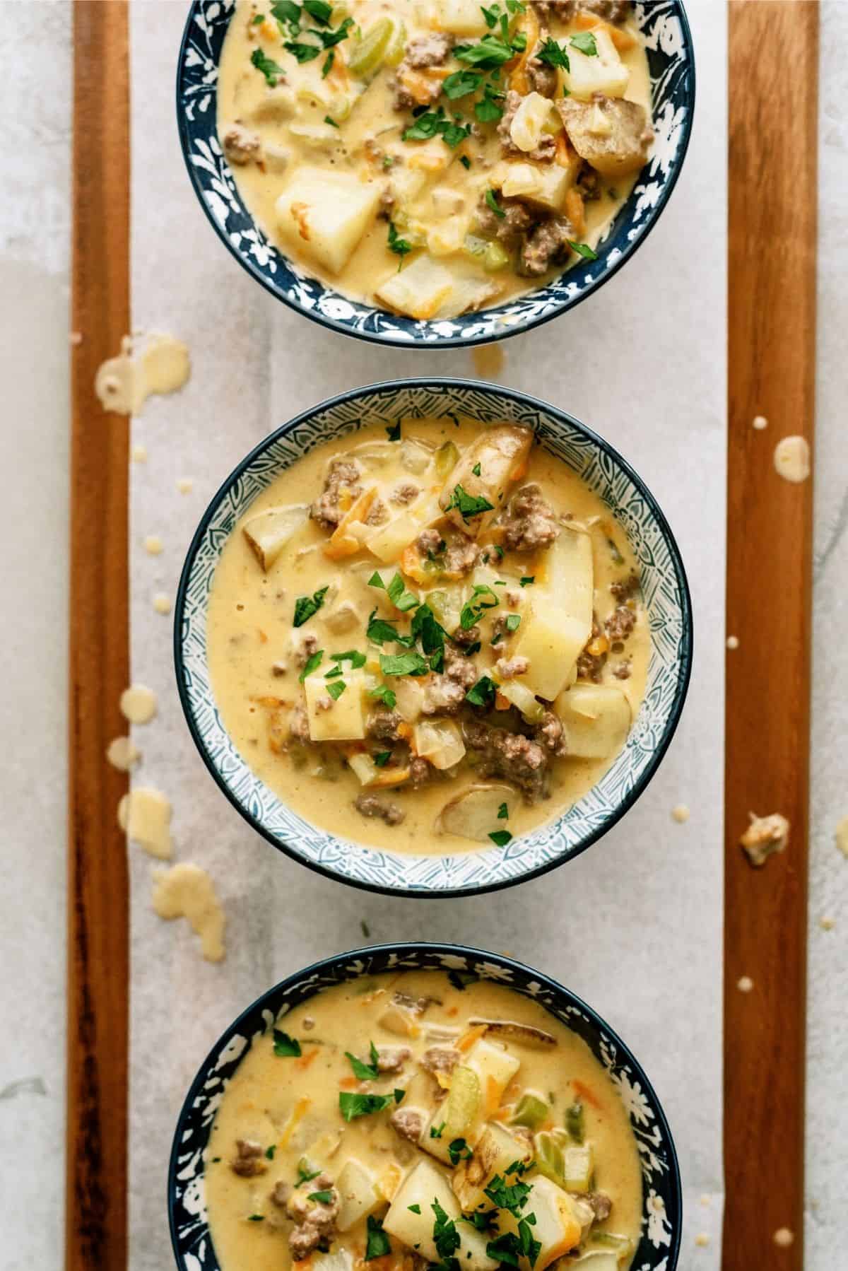 3 Bowls full of The Best Cheeseburger Soup Recipe (Cheesy Ground Beef Soup) on a wooden board