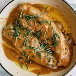 Cooked applesauce pork tenderloin in a light-colored pan, covered with brown sauce and garnished with fresh rosemary sprigs.