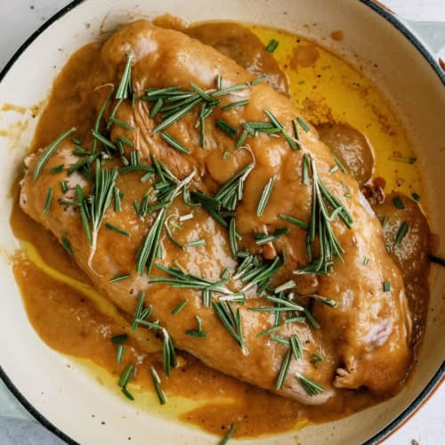 Cooked applesauce pork tenderloin in a light-colored pan, covered with brown sauce and garnished with fresh rosemary sprigs.