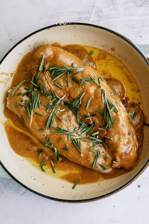 Cooked applesauce pork tenderloin in a light-colored pan, covered with brown sauce and garnished with fresh rosemary sprigs.