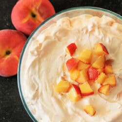 A glass bowl filled with creamy dessert topped with diced peaches, placed next to two whole peaches on a dark surface.