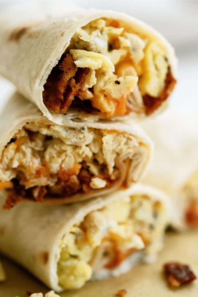 Three halved breakfast burritos stacked, showing fillings of scrambled eggs, cheese, and sausage inside flour tortillas.
