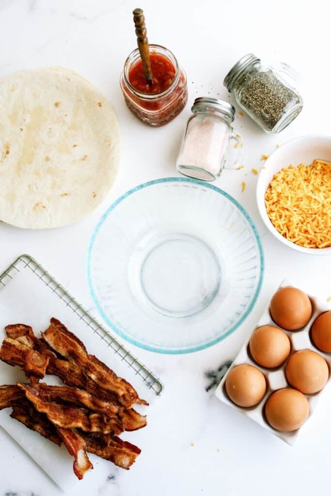Ingredients for a breakfast wrap include a tortilla, cooked bacon, eggs, shredded cheese, salt, pepper, and salsa, arranged on a white surface with a glass bowl in the center.