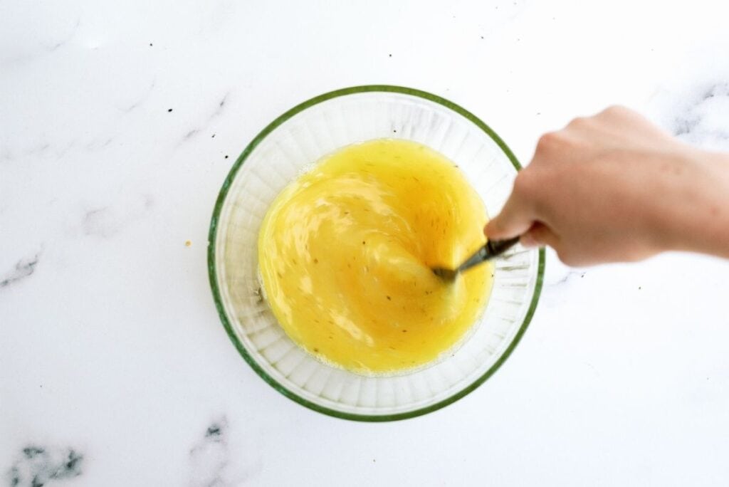 A hand whisking yellow liquid in a glass bowl on a white countertop.