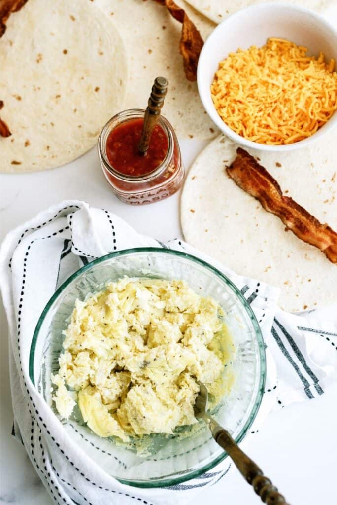 A bowl of scrambled eggs, sliced bacon, shredded cheese, tortillas, and a jar of salsa on a white cloth.