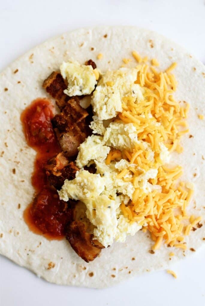 A tortilla topped with scrambled eggs, shredded cheese, pieces of bacon, and salsa on a white background.