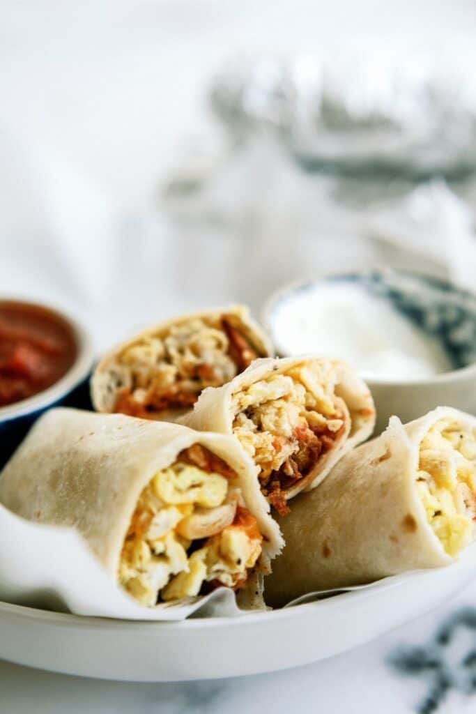 Close-up of a plate with three breakfast burrito halves filled with scrambled eggs and meat. A small bowl of salsa and a bowl of sour cream are in the background.